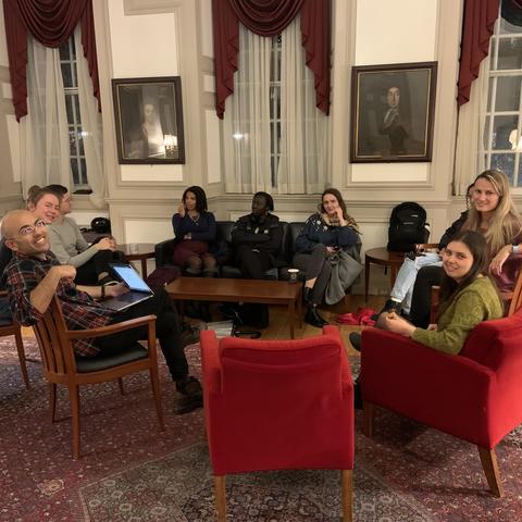 Students sit in circle in comfy armchairs for a discussion. Many look towards the camera for the picture. 