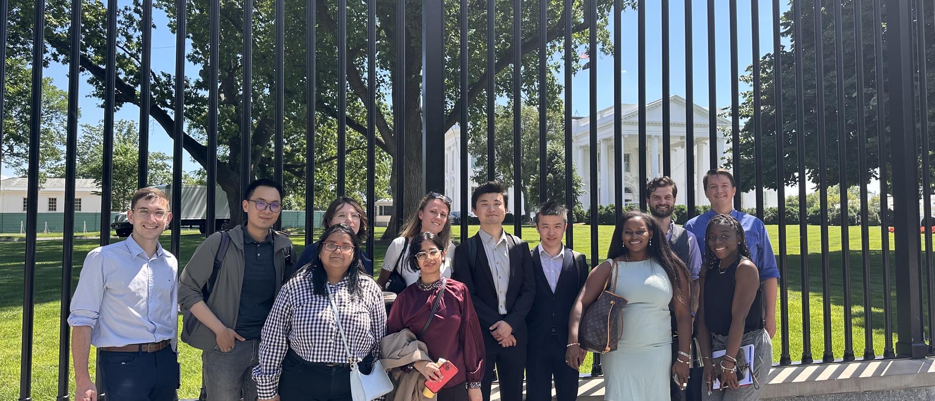 A group of well dressed students stand in front of the fence in front of the White House on a sunny day